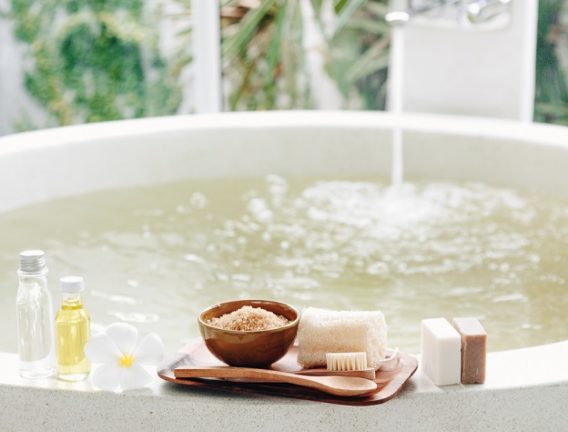 Lifestyle image of a tray on the edge of a bath, holding a sponge, bathing brush and bath salts, as well bars of soap and bottles of body wash